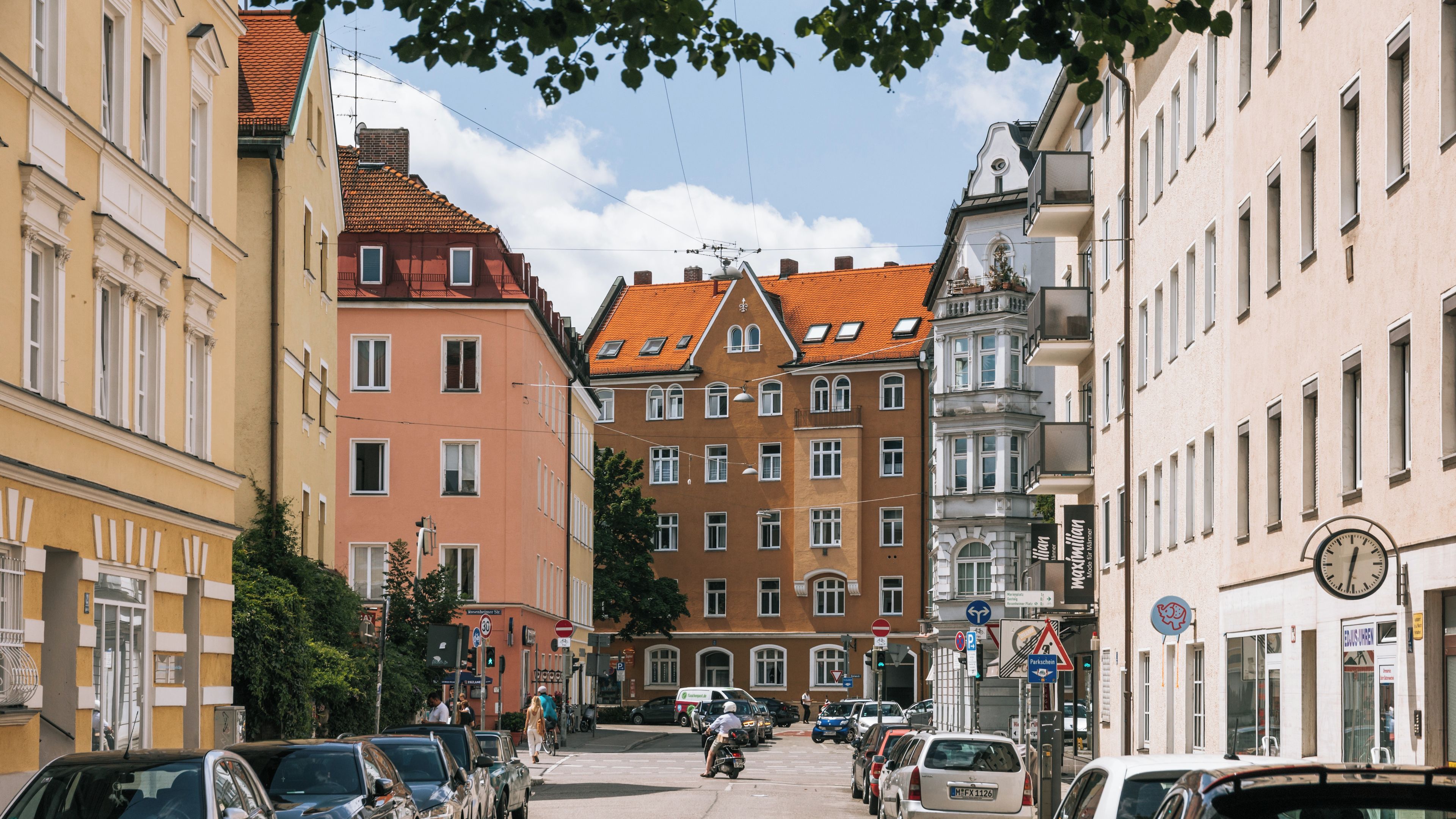 A European street lined with historic buildings, parked cars, and a cyclist.