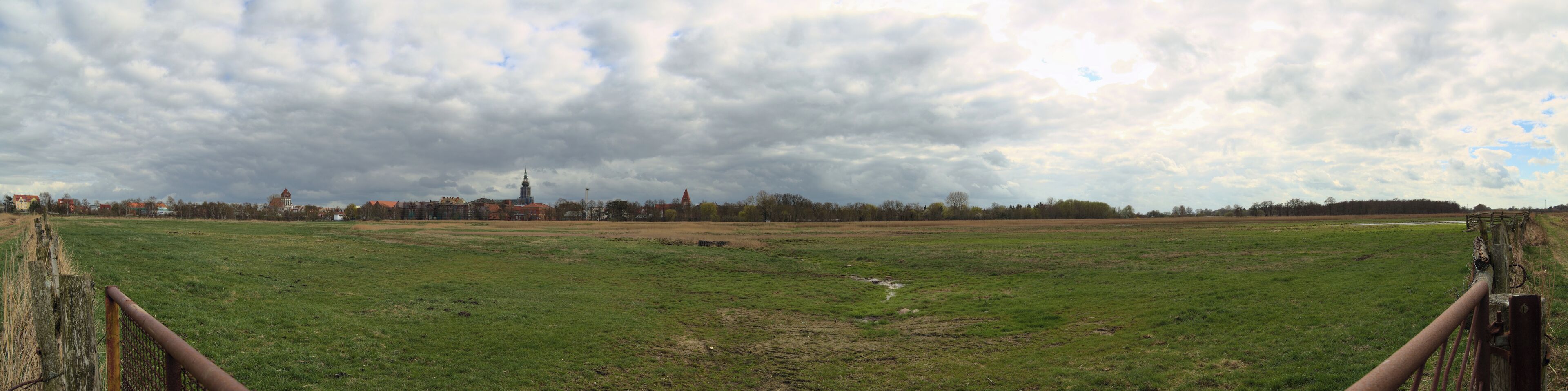 A rural landscape with a grassy field, a wooden fence, and a distant town with buildings and a tower.
