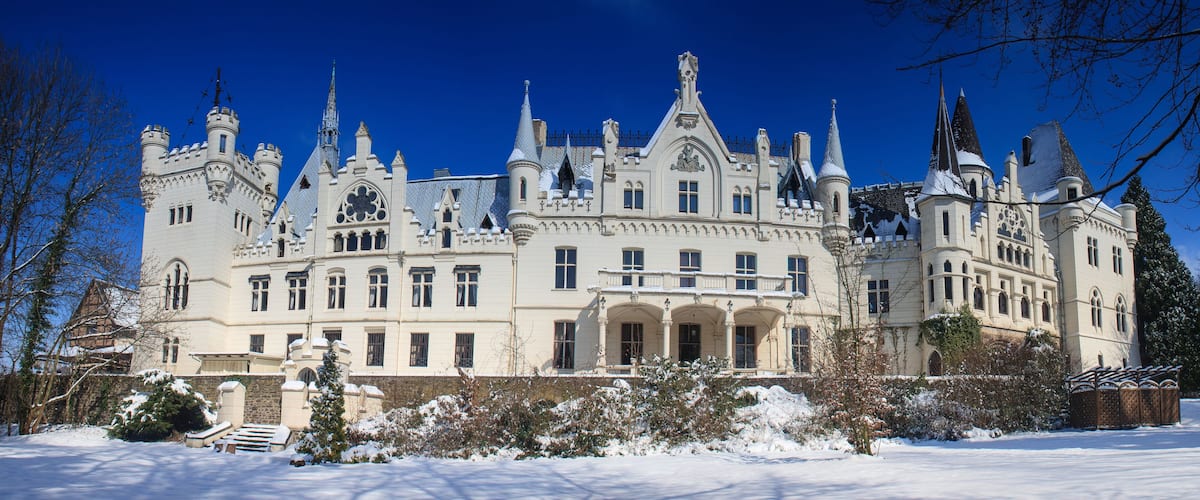 Ein großes, verziertes Schloss mit zahlreichen Türmen und Zinnen, umgeben von schneebedeckten Bäumen und blauem Himmel.