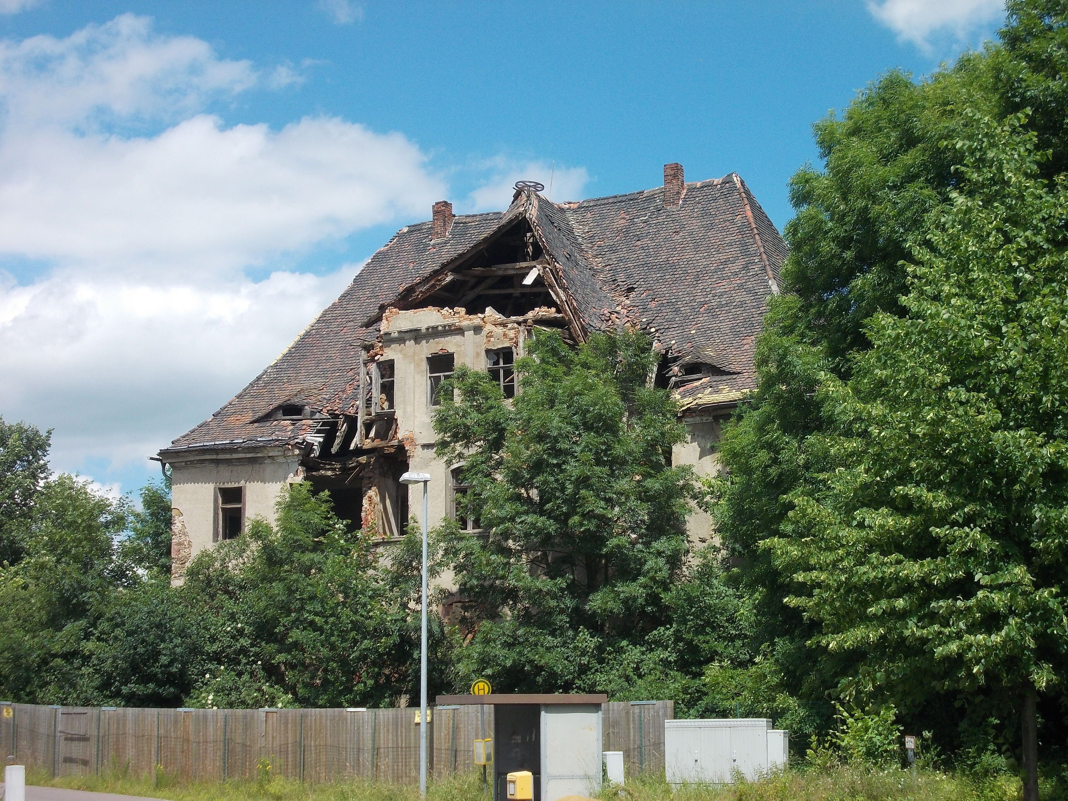 A dilapidated building with a damaged roof and overgrown vegetation.