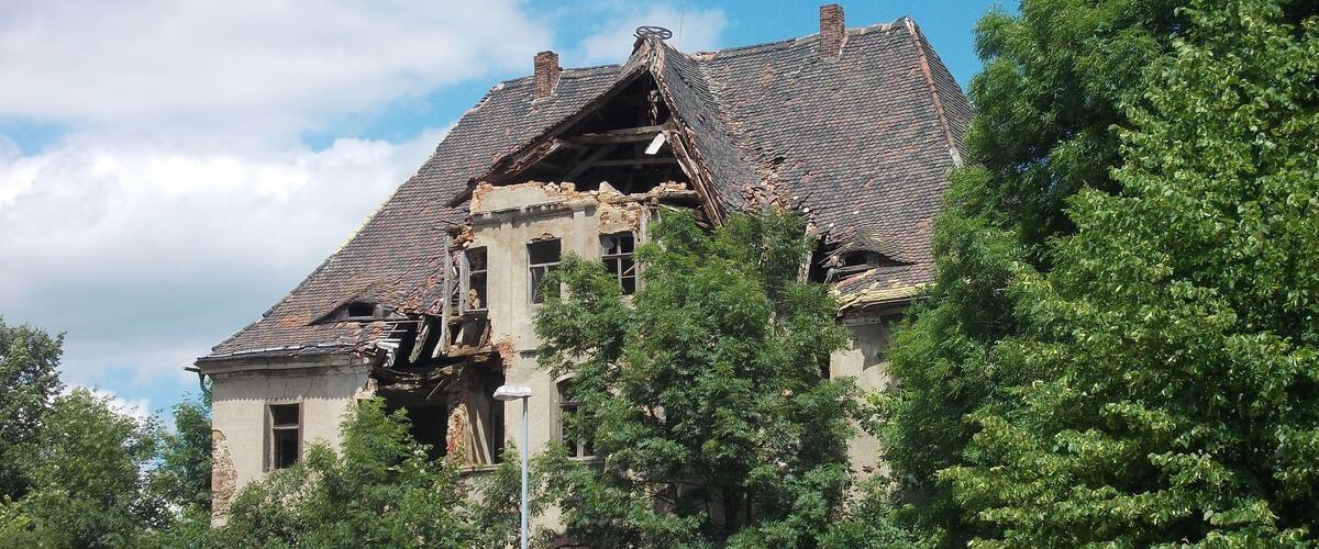 A dilapidated building with a damaged roof and overgrown vegetation.