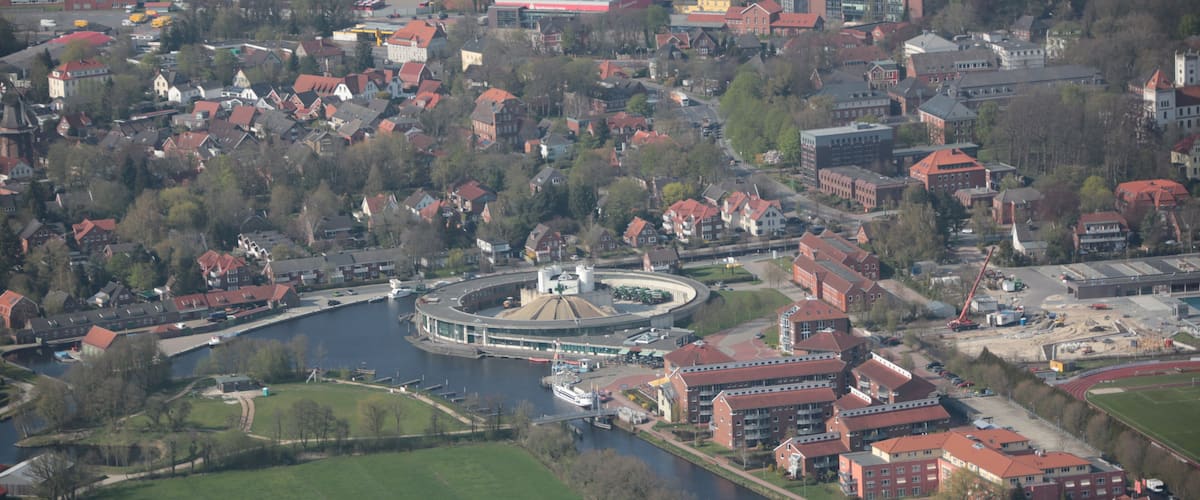 An aerial view of a town with a river, green fields, and red-roofed buildings.