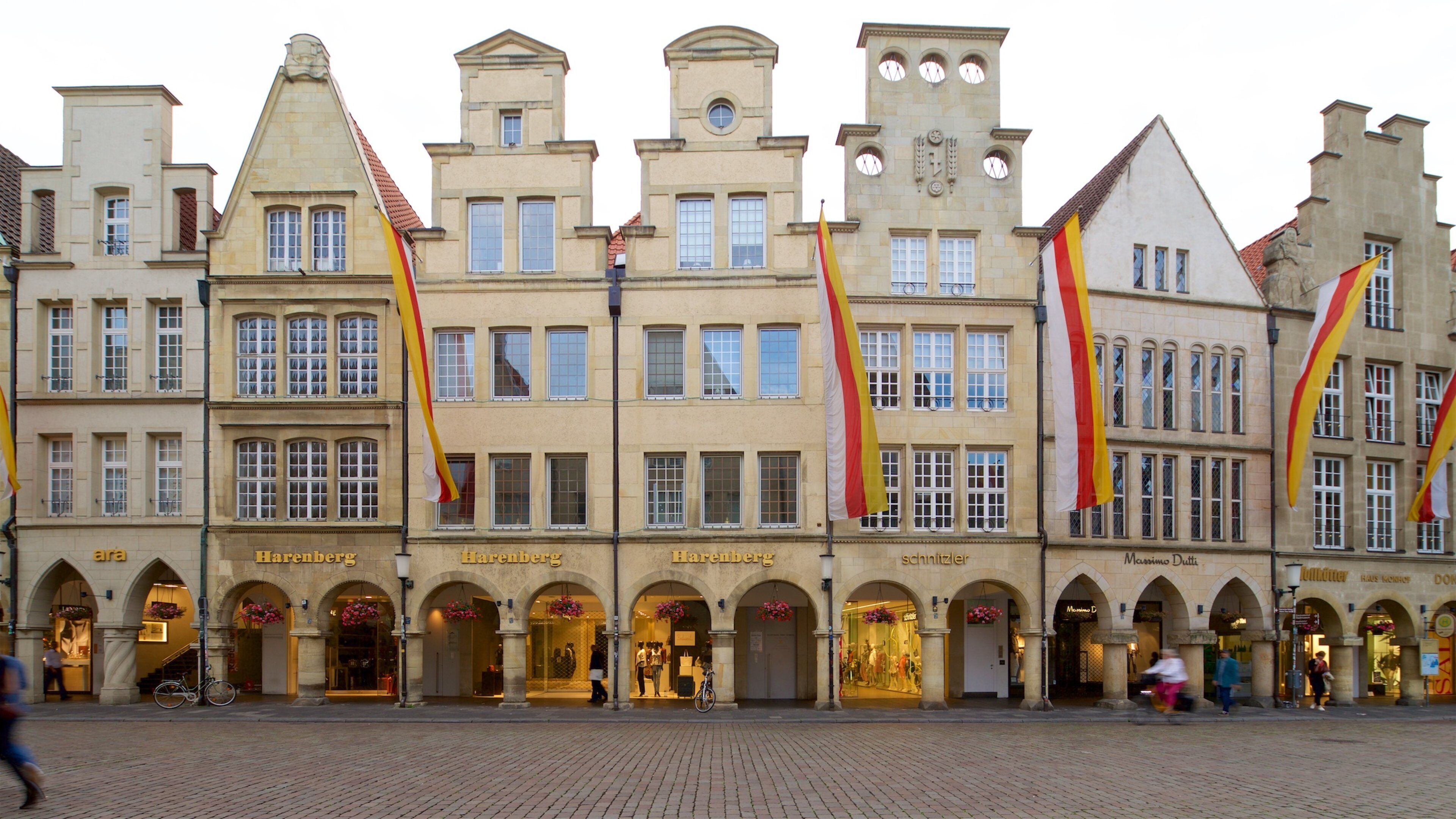 A historic European street with multi-story buildings, prominent flags, and storefronts.