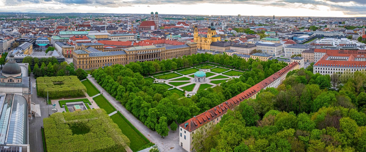 Eine Stadtansicht mit einem markanten Platz, der von üppigen Grünflächen und historischen Gebäuden umgeben ist und ein zentrales Denkmal beherbergt.