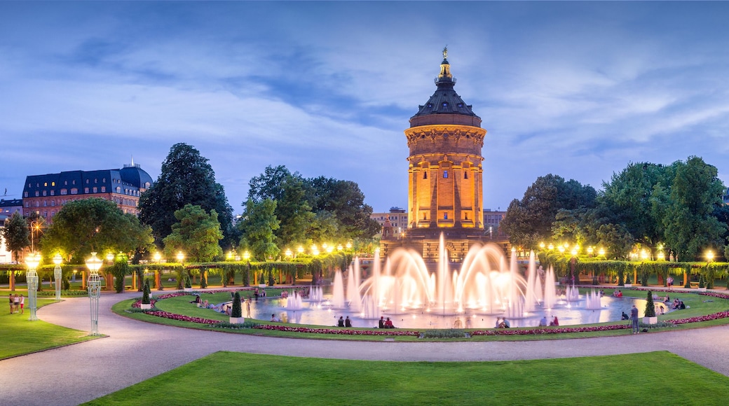A historic tower with a spire, surrounded by a well-lit fountain and manicured gardens.
