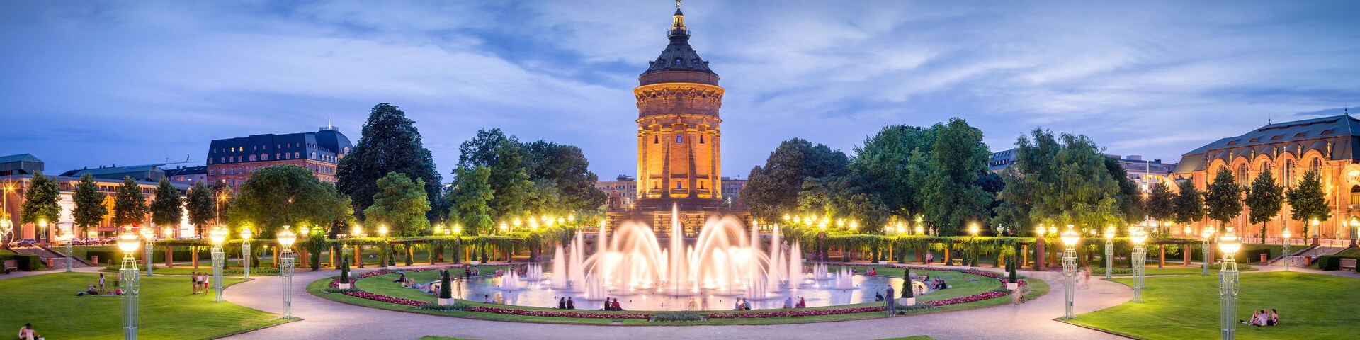 A historic tower with a spire, surrounded by a well-lit fountain and manicured gardens.