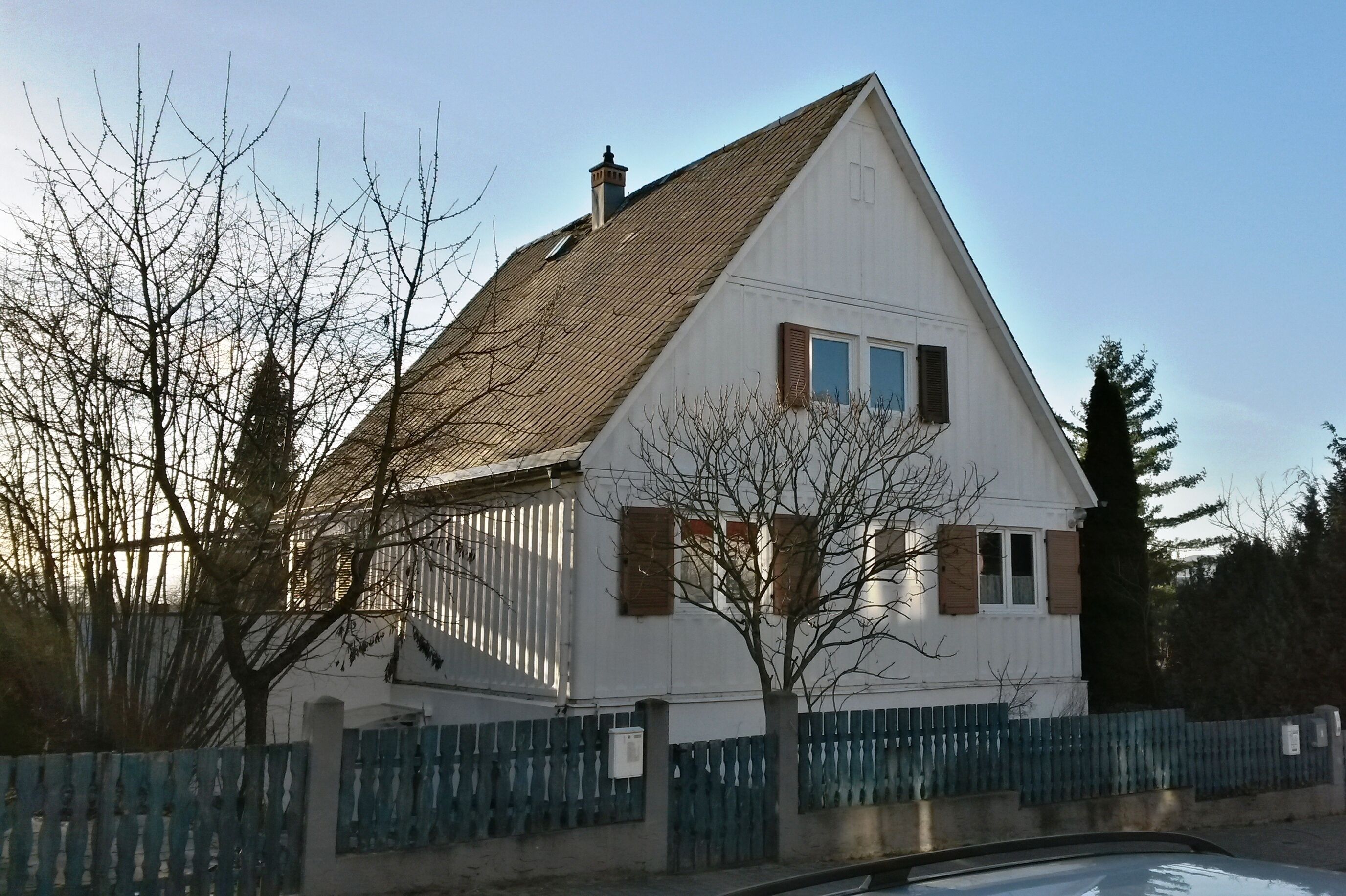 A white house with a brown roof, surrounded by a blue fence and bare trees.