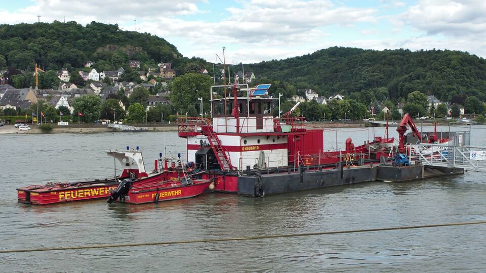 A red and white tugboat named 'Feuerwehr' docked at a harbor with a backdrop of a hill and houses.