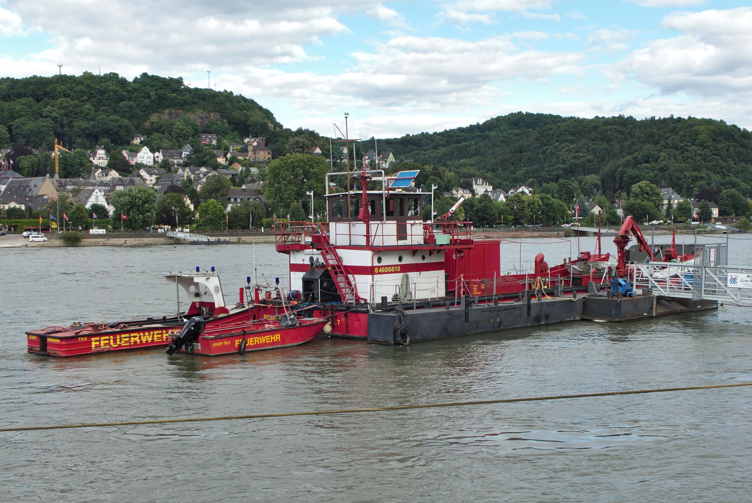 A red and white tugboat named 'Feuerwehr' docked at a harbor with a backdrop of a hill and houses.