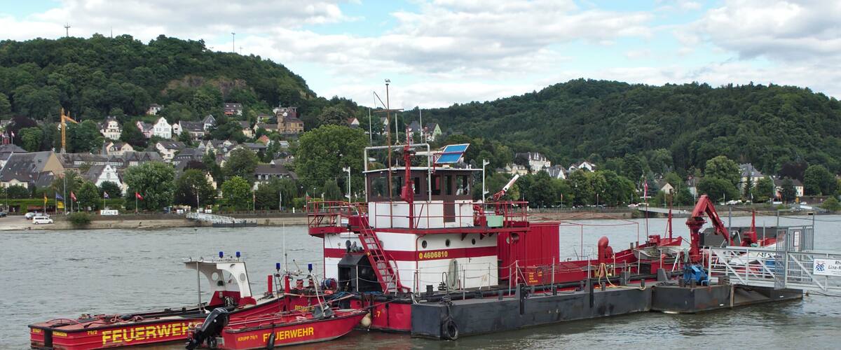 A red and white tugboat named 'Feuerwehr' docked at a harbor with a backdrop of a hill and houses.