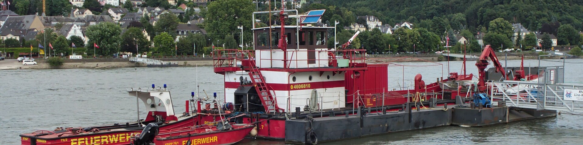 A red and white tugboat named 'Feuerwehr' docked at a harbor with a backdrop of a hill and houses.