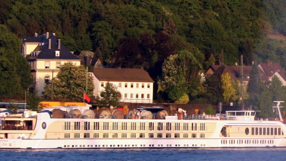 A river cruise ship with multiple decks, surrounded by a forested hillside and residential buildings.