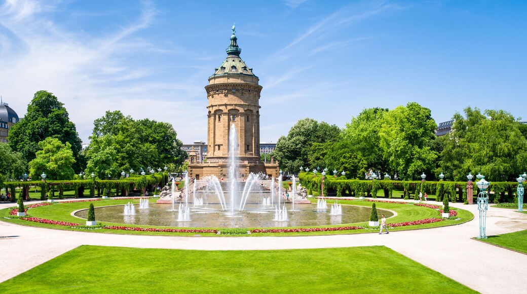 A historic tower with a statue on top, surrounded by a well-maintained garden with fountains and neatly trimmed hedges.