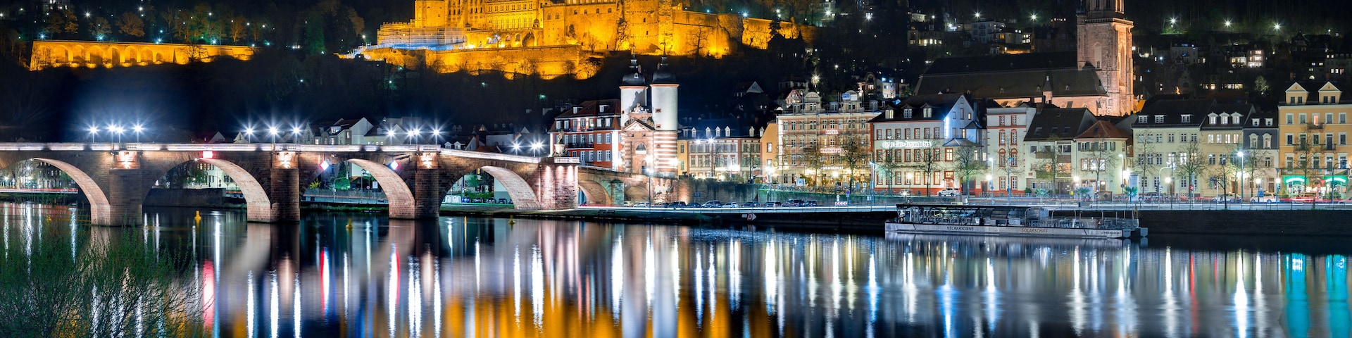 Una vista nocturna de una ciudad con un castillo histórico iluminado sobre una colina, un puente cruzando un río y un barco amarrado a la orilla del río.