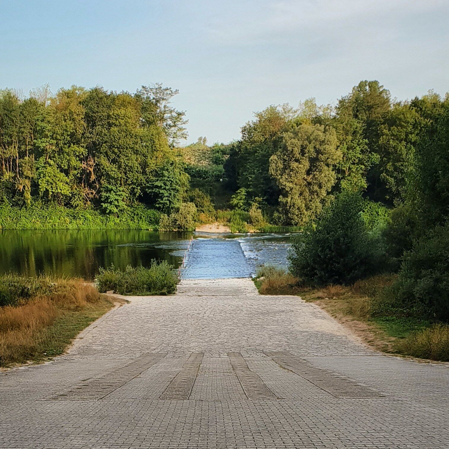 A paved pathway leading to a calm river surrounded by dense greenery.
