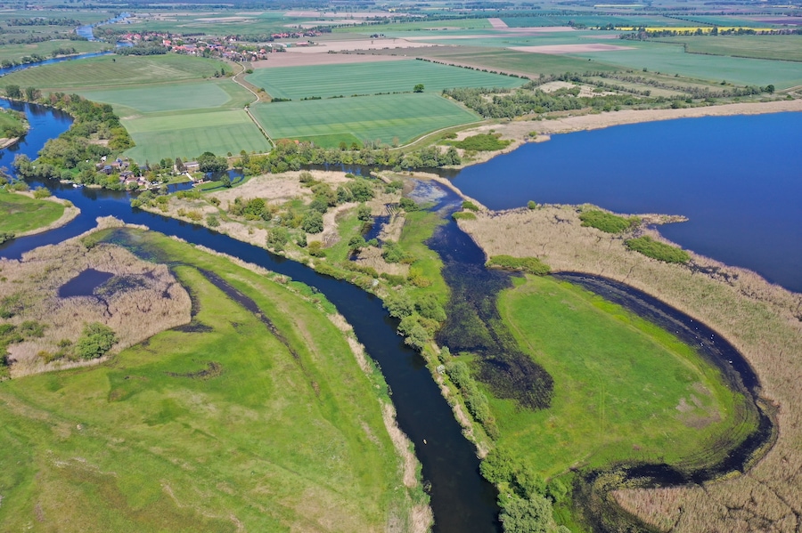 Vista aérea de un río serpenteando a través de campos verdes, con un pequeño asentamiento y una gran masa de agua a la derecha.