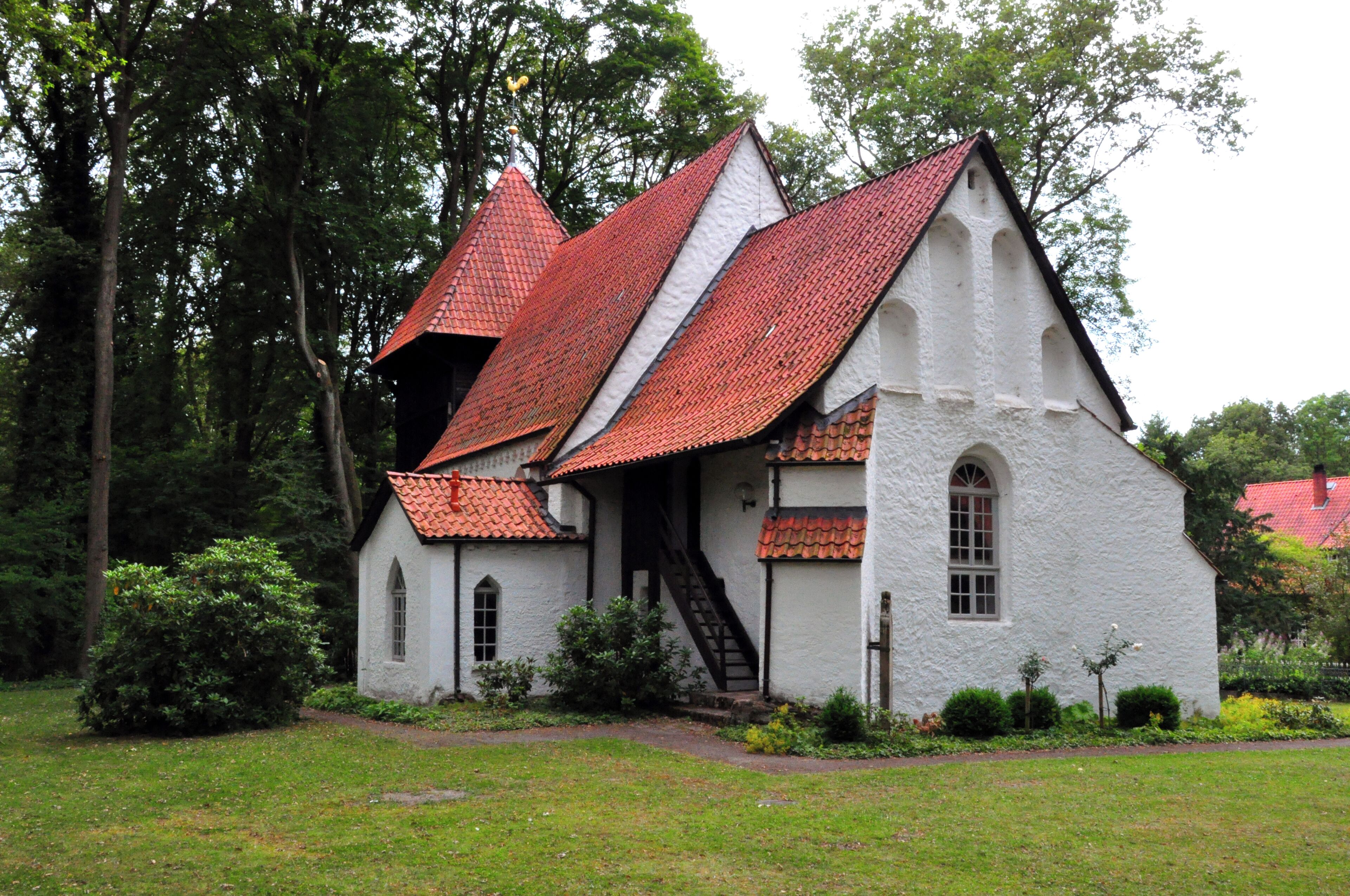 A white church with red-tiled roofs and a small bell tower, surrounded by lush greenery.