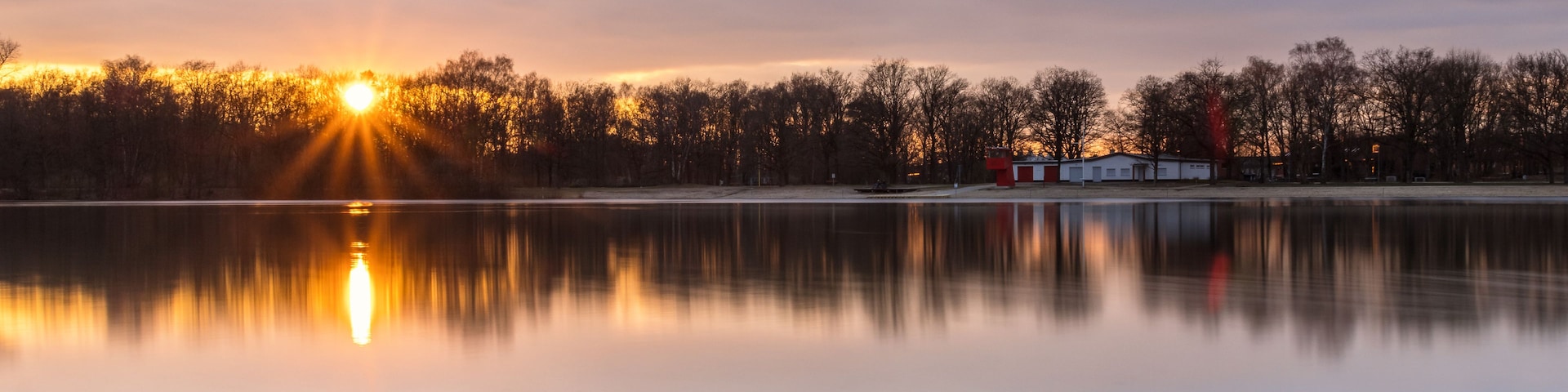 Un lago tranquilo al atardecer, con el sol reflejándose en el agua, rodeado de árboles y un pequeño edificio con techo rojo.