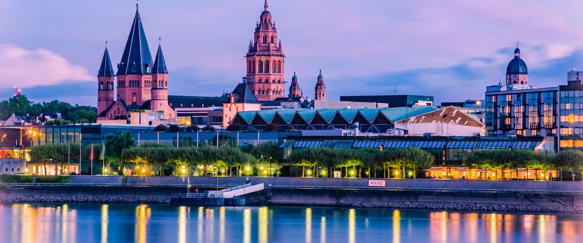 Eine Stadtansicht mit einem Fluss im Vordergrund, zeigt historische Gebäude mit Spitzentürmen und einer Kuppelkonstruktion.