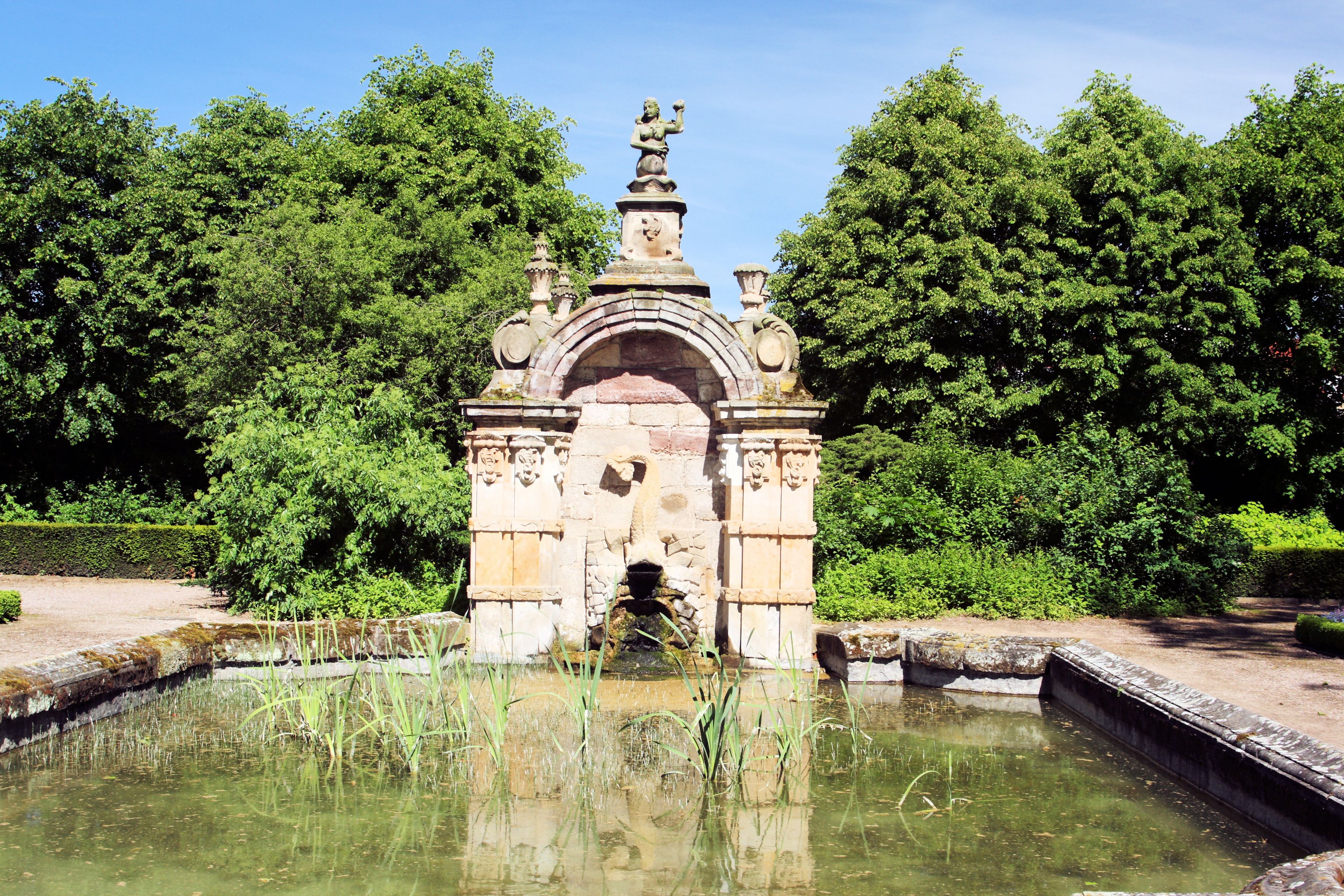 A classical stone fountain with a statue on top, surrounded by lush greenery.