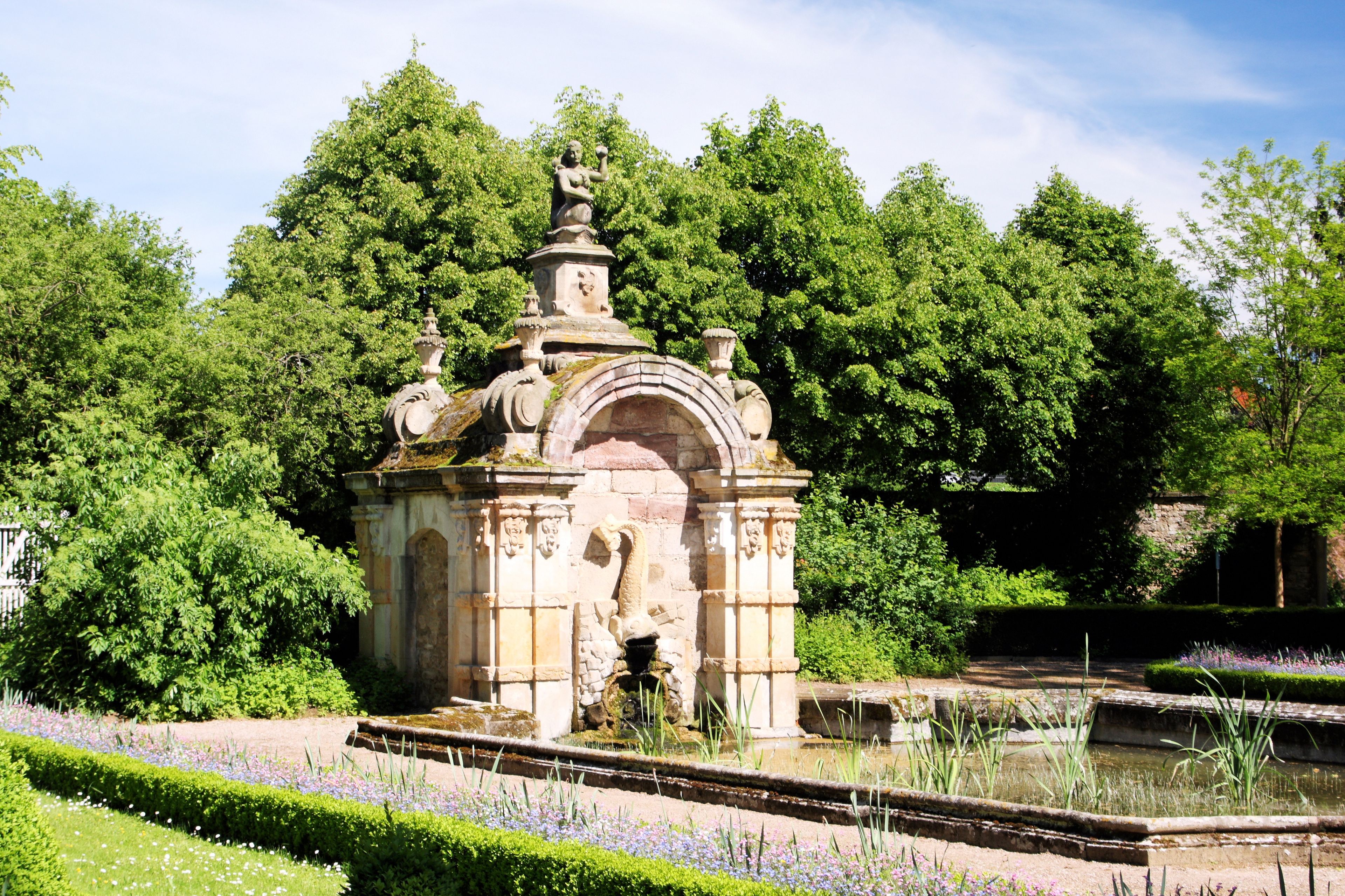 A classical stone fountain with intricate carvings, surrounded by lush greenery and a well-maintained garden with flowering plants.