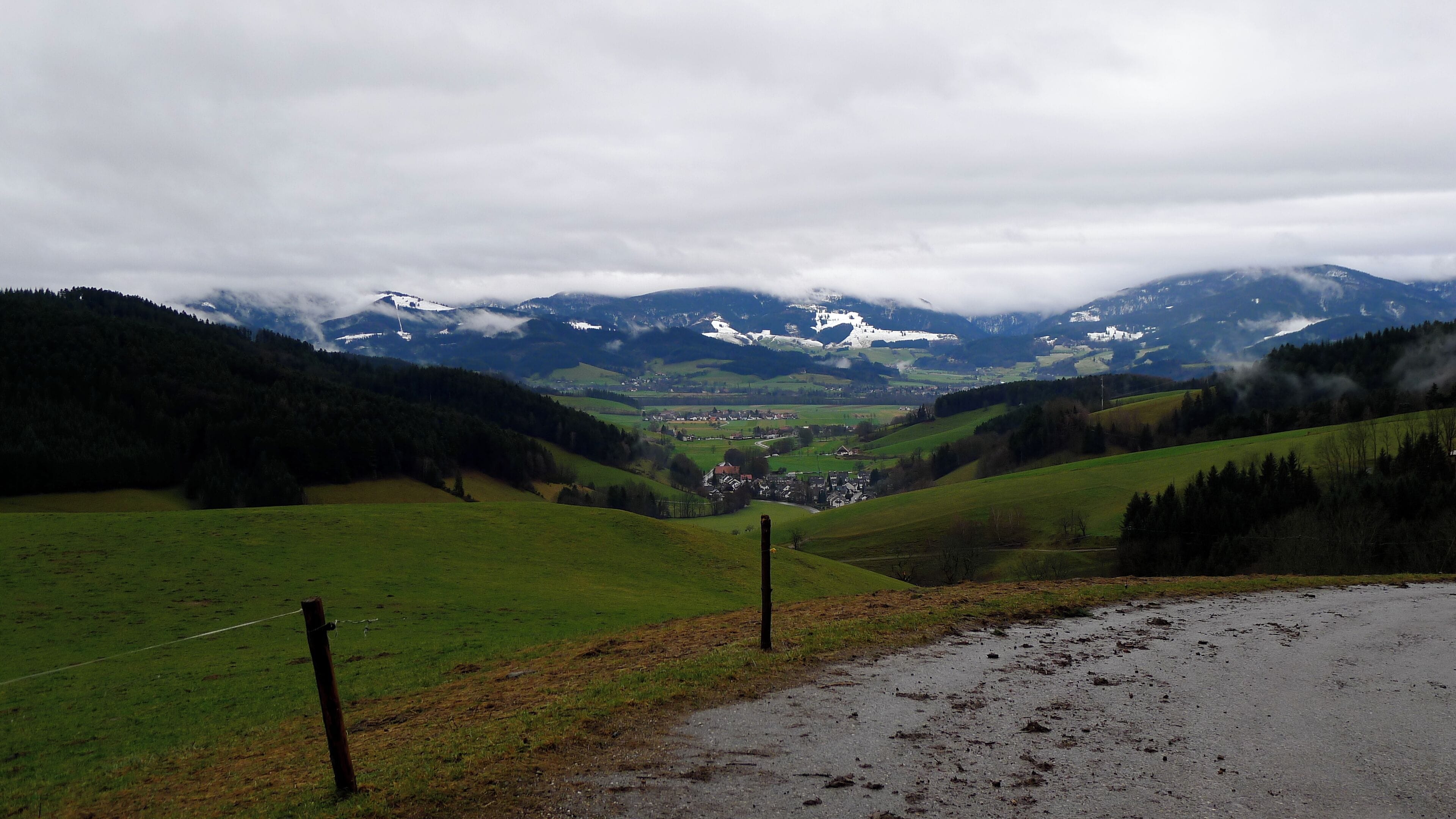 A valley with a road, green fields, and distant snow-capped mountains under a cloudy sky.