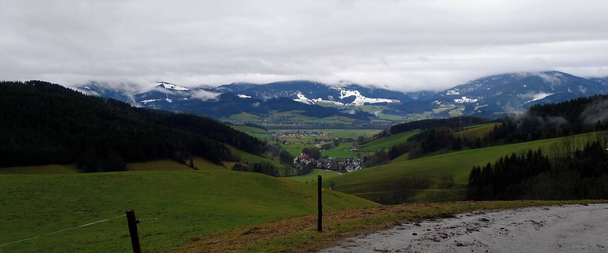 A valley with a road, green fields, and distant snow-capped mountains under a cloudy sky.