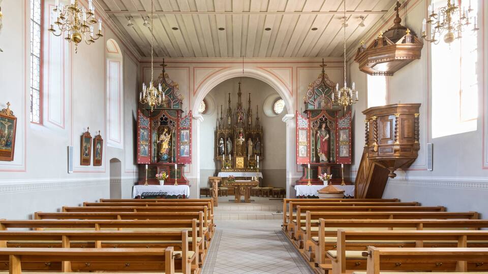 An interior of a church with wooden pews, a central altar, and ornate chandeliers.