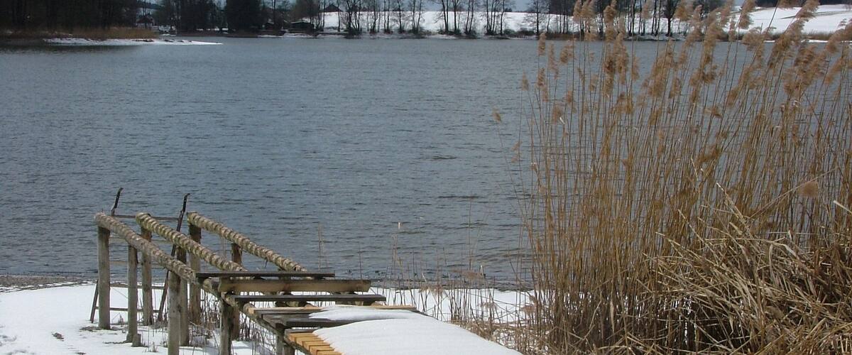 A wooden bench with snow on it, a frozen lake, and bare trees in the background.