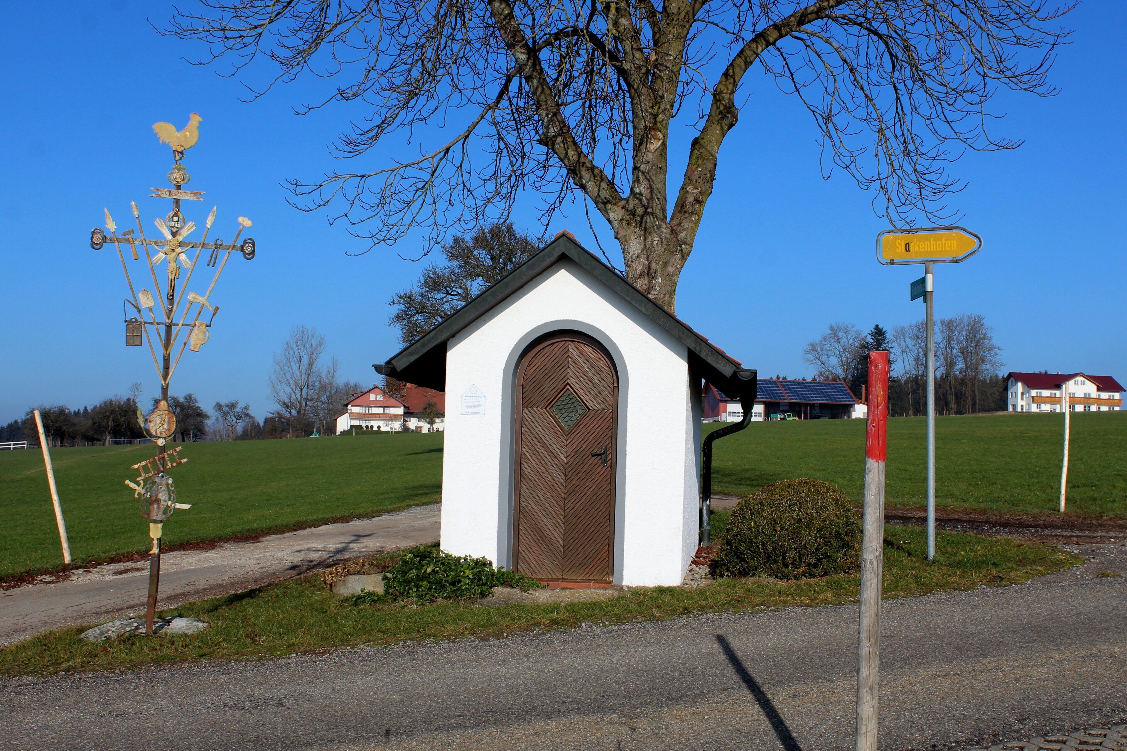 A small white chapel with a brown door, a weather vane, and a directional sign.