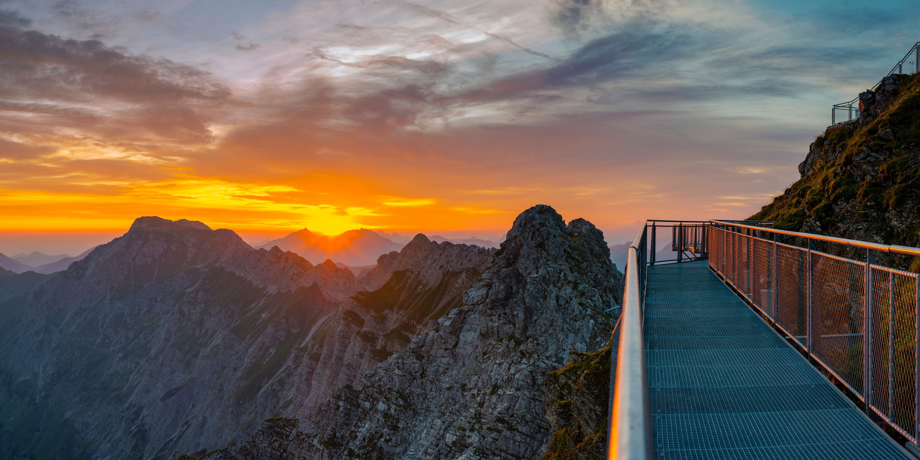 A metal walkway with a metal railing, overlooking a mountainous landscape during sunset.