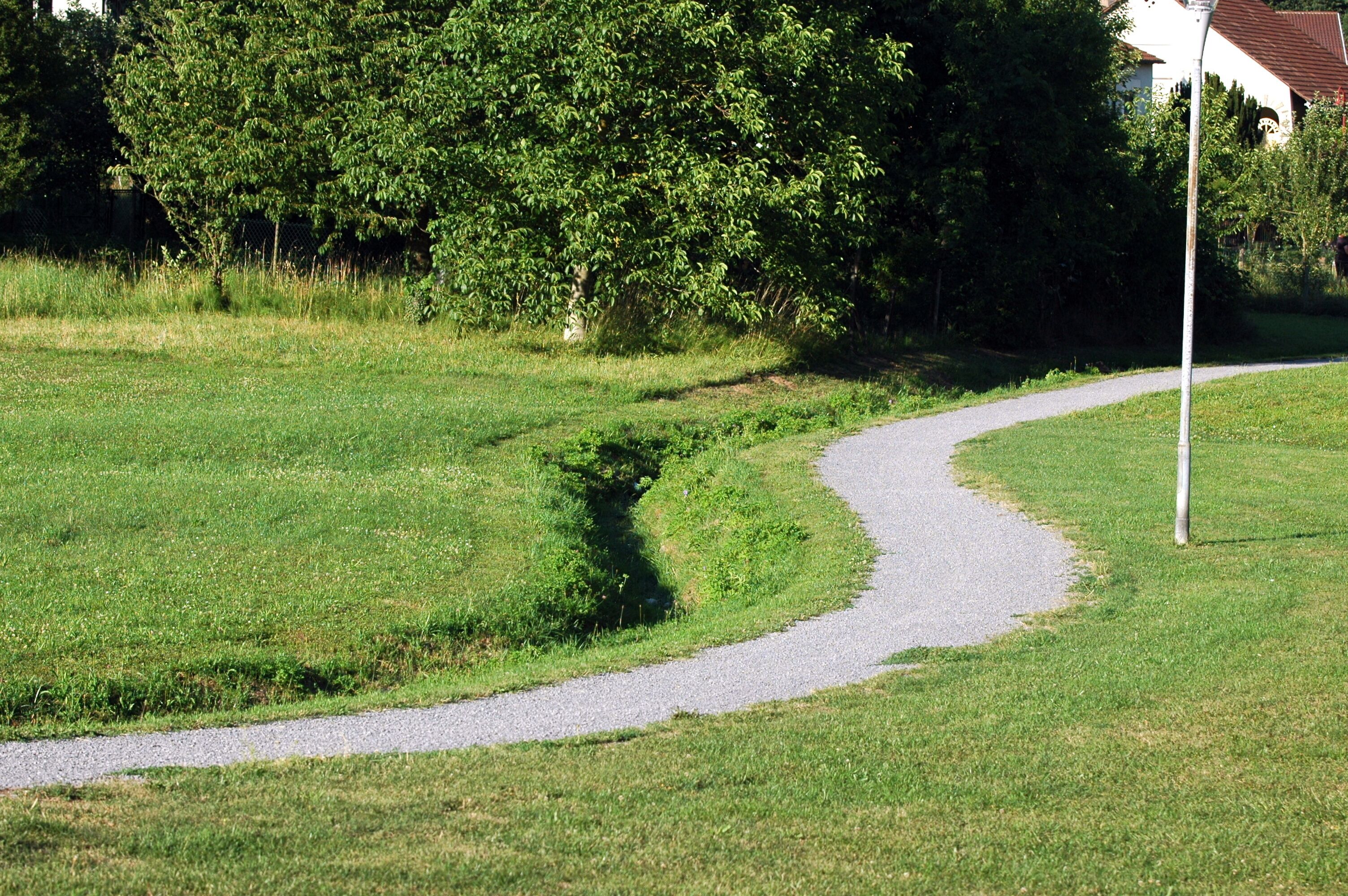 A winding gravel path in a grassy area with trees and a building in the background.