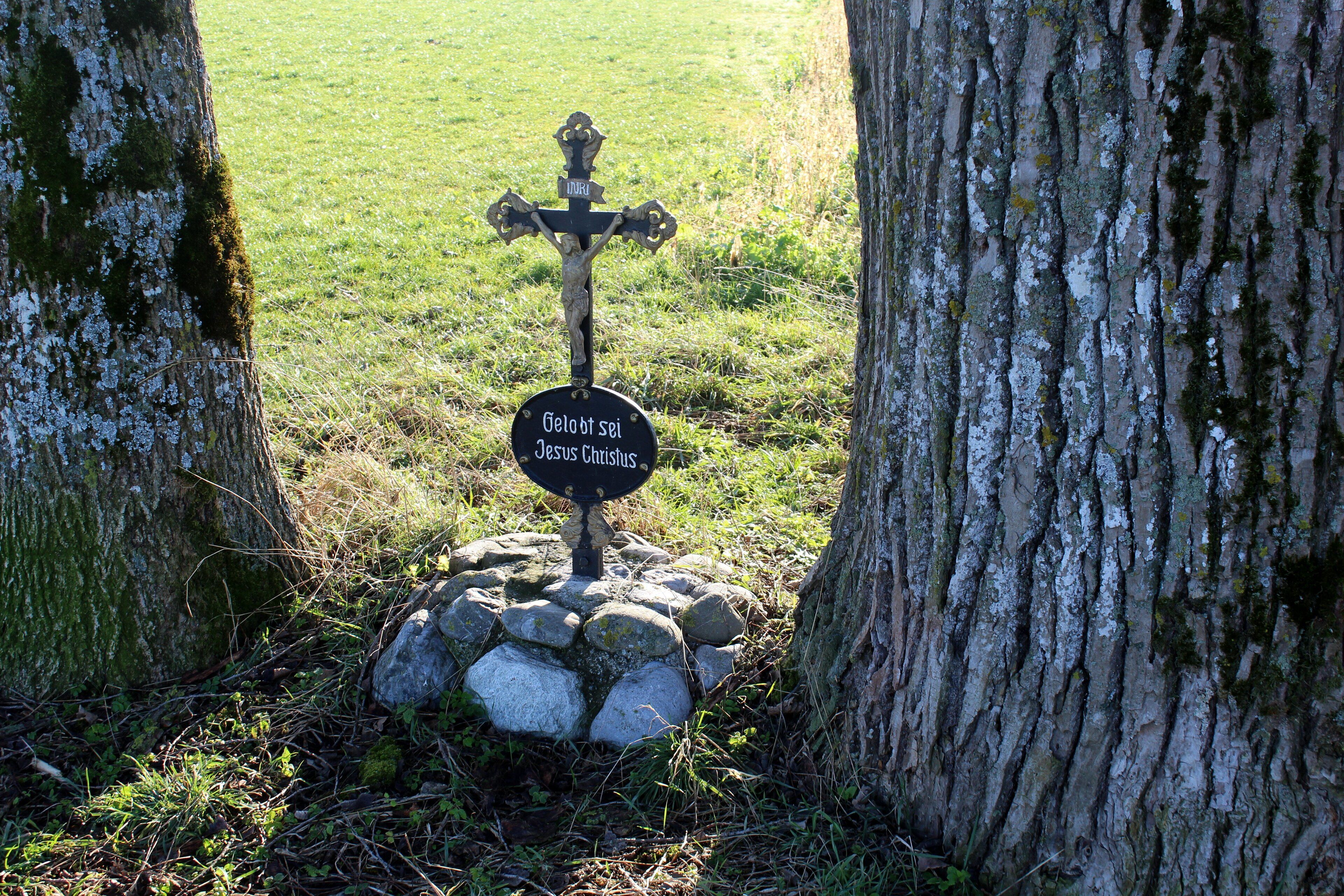 A stone cross with a sign reading "Geloft Sel Jesus Christus" surrounded by rocks and trees.