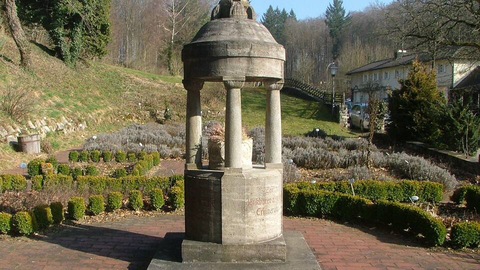 A stone monument with inscriptions, surrounded by a landscaped garden with hedges and trees.