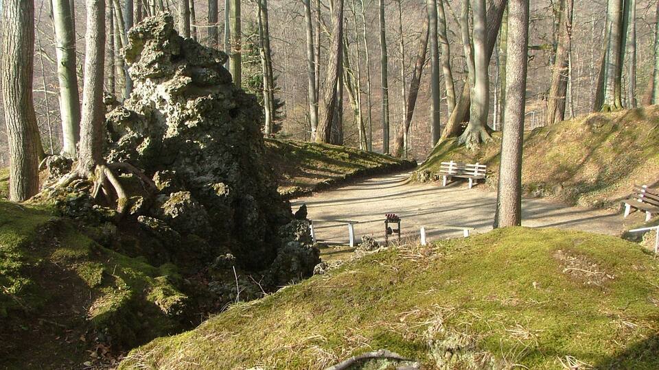 A forested area with a dirt path, a large moss-covered rock, and benches along the path.