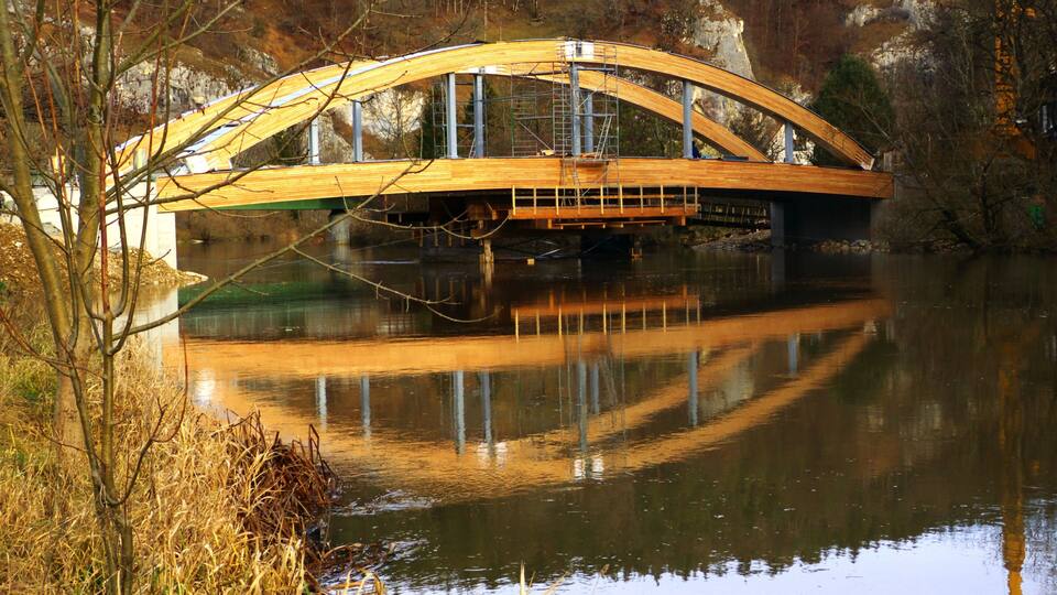 A wooden bridge over a calm river, surrounded by trees and rocky terrain.