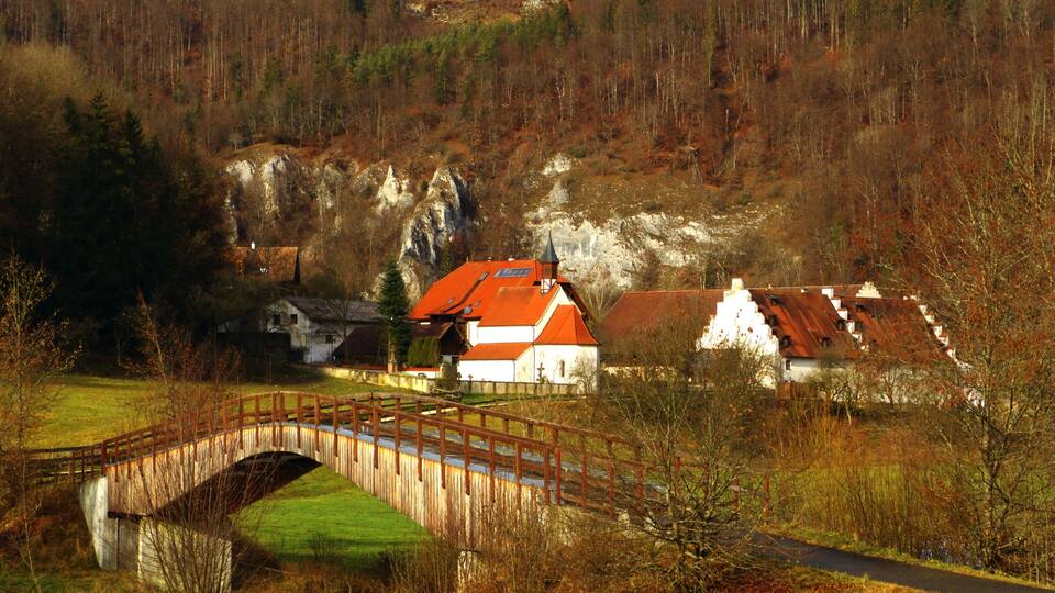 A rural landscape with a wooden bridge, a river, and houses with red roofs.