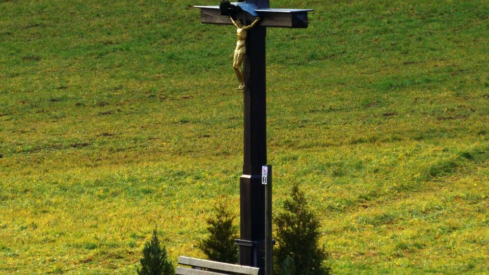 A wooden cross with a figure, a bench, and a small tree in a grassy field.