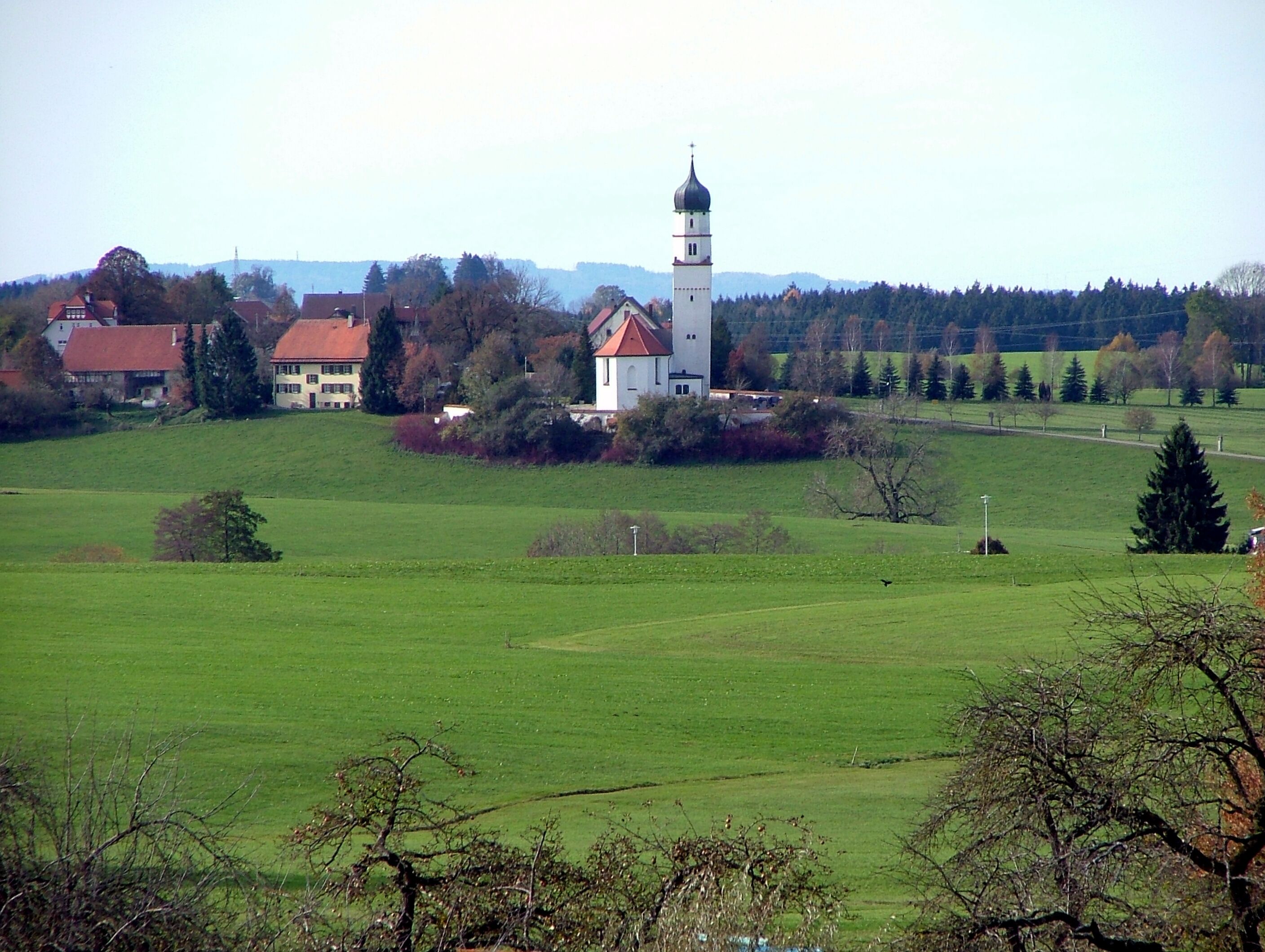 A church with a tall tower and a red roof, surrounded by green fields and trees.