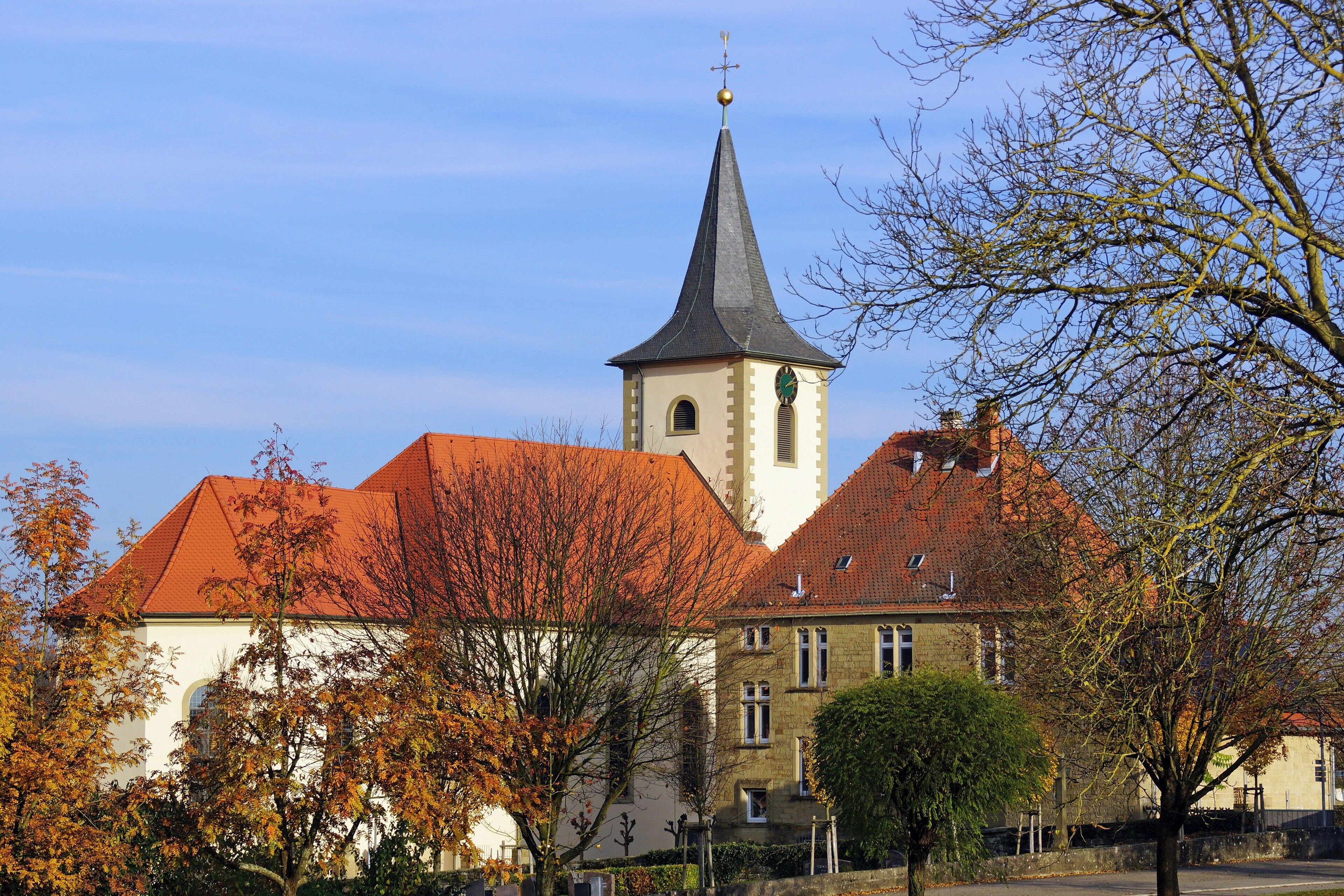 A church with a tall steeple and a clock tower, surrounded by autumn trees and buildings with red roofs.