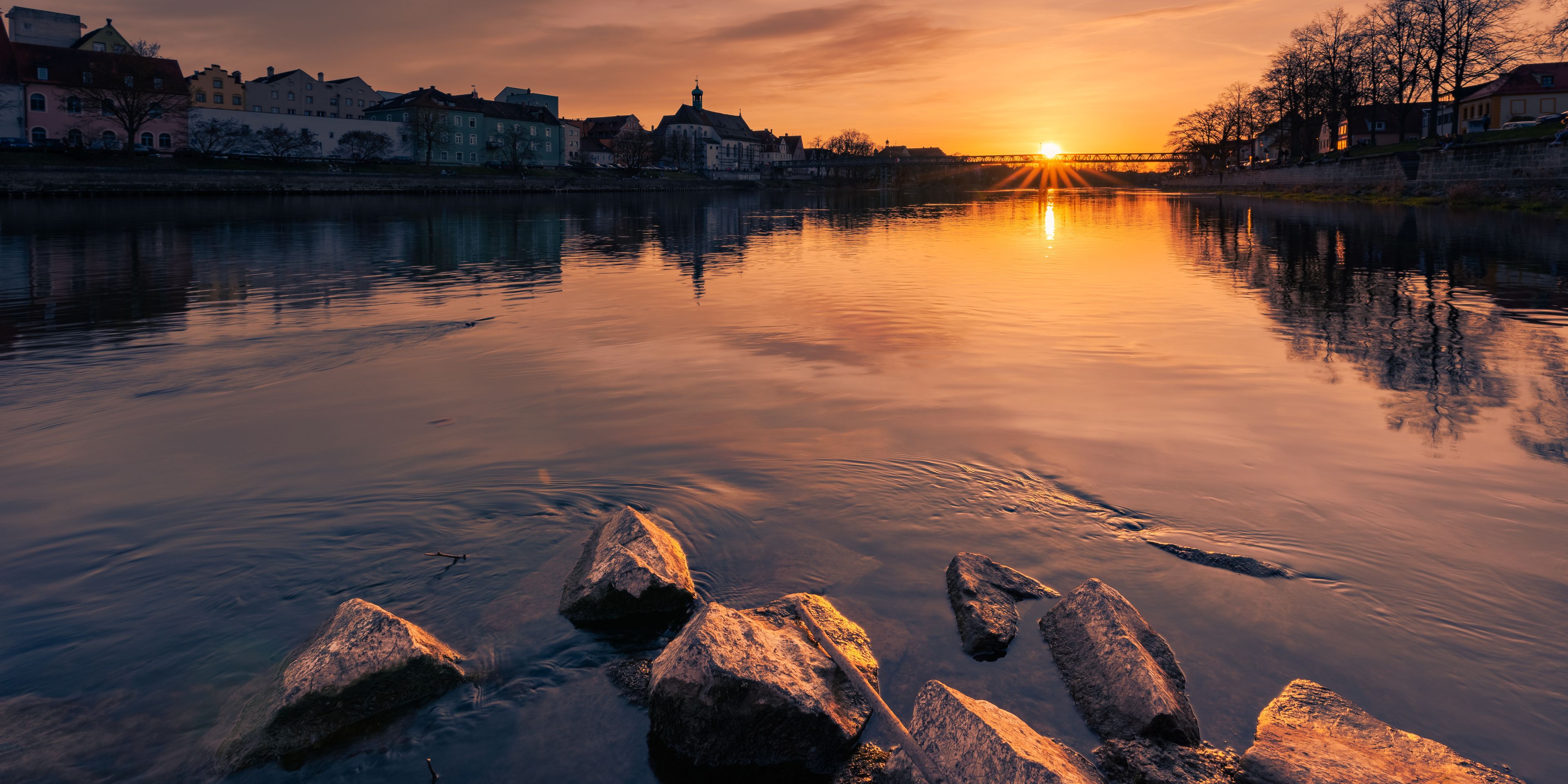 Un atardecer sobre un río tranquilo, con edificios a la otra orilla y un puente en la distancia.