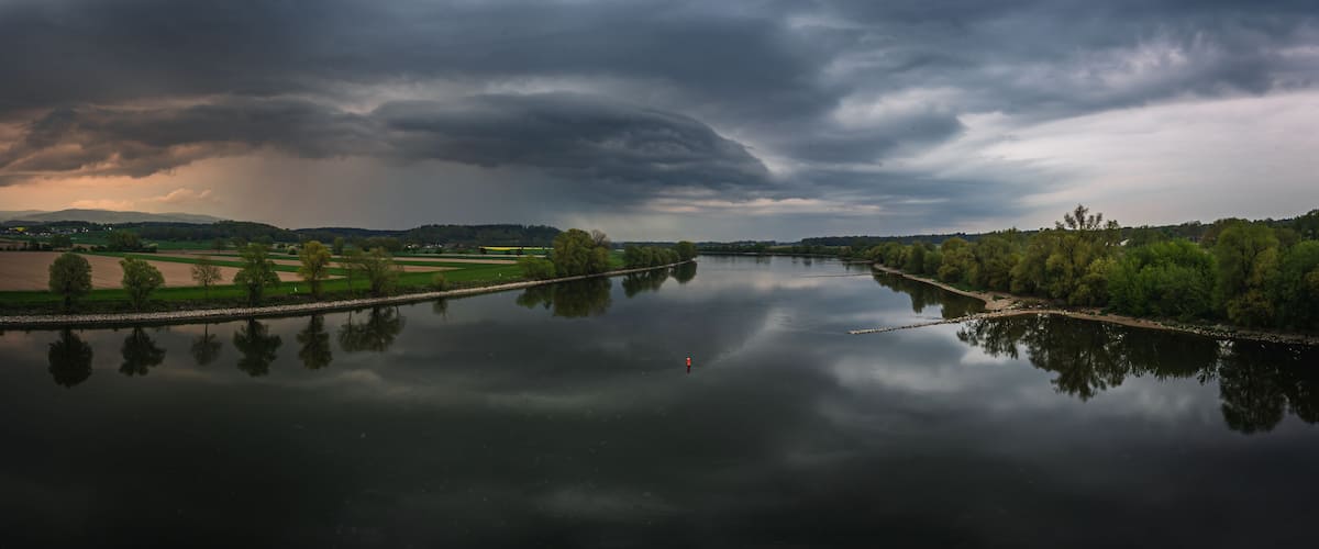 A river reflecting a stormy sky, surrounded by fields and trees.
