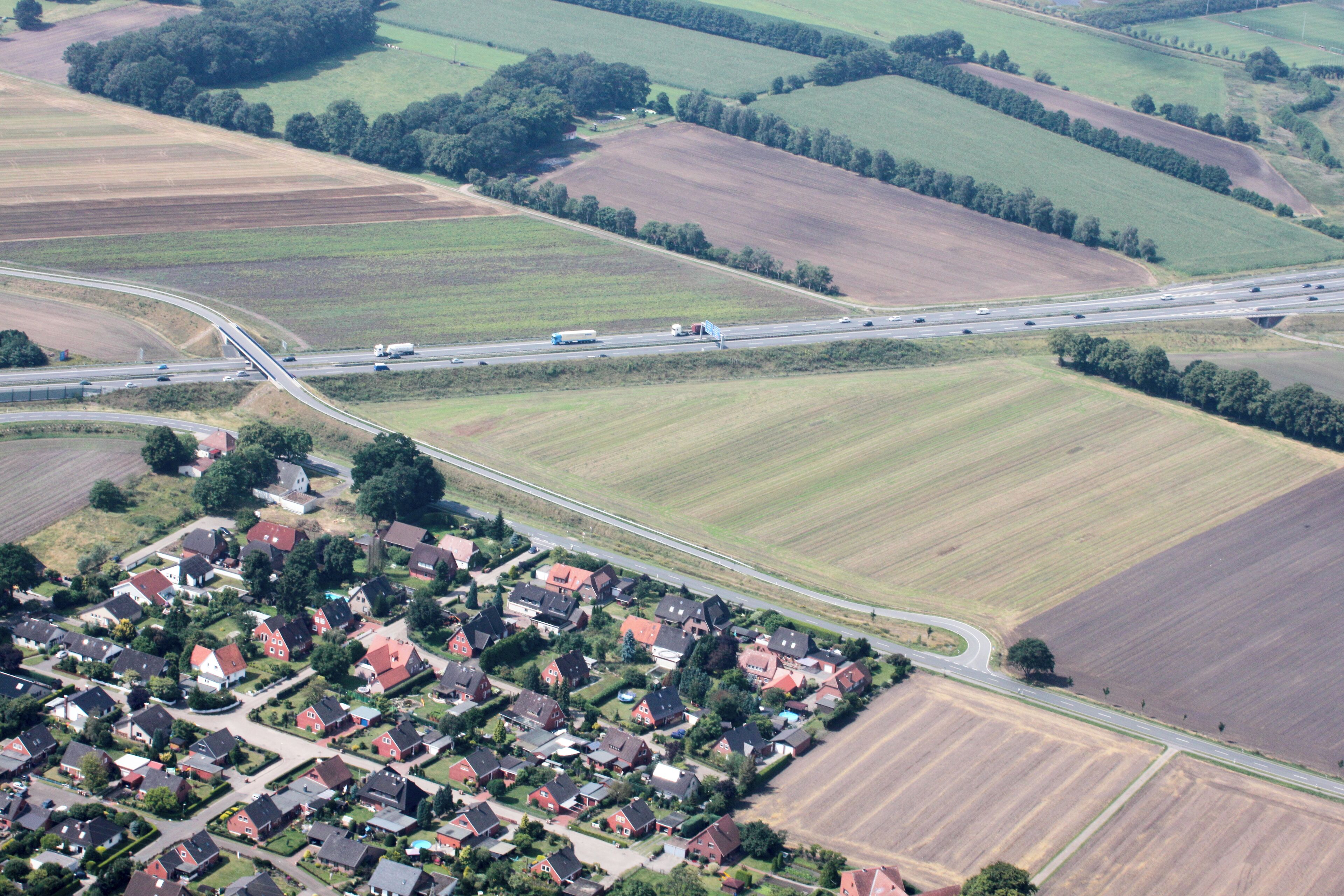 An aerial view of a rural area with a highway, fields, and a residential area with red-roofed houses.