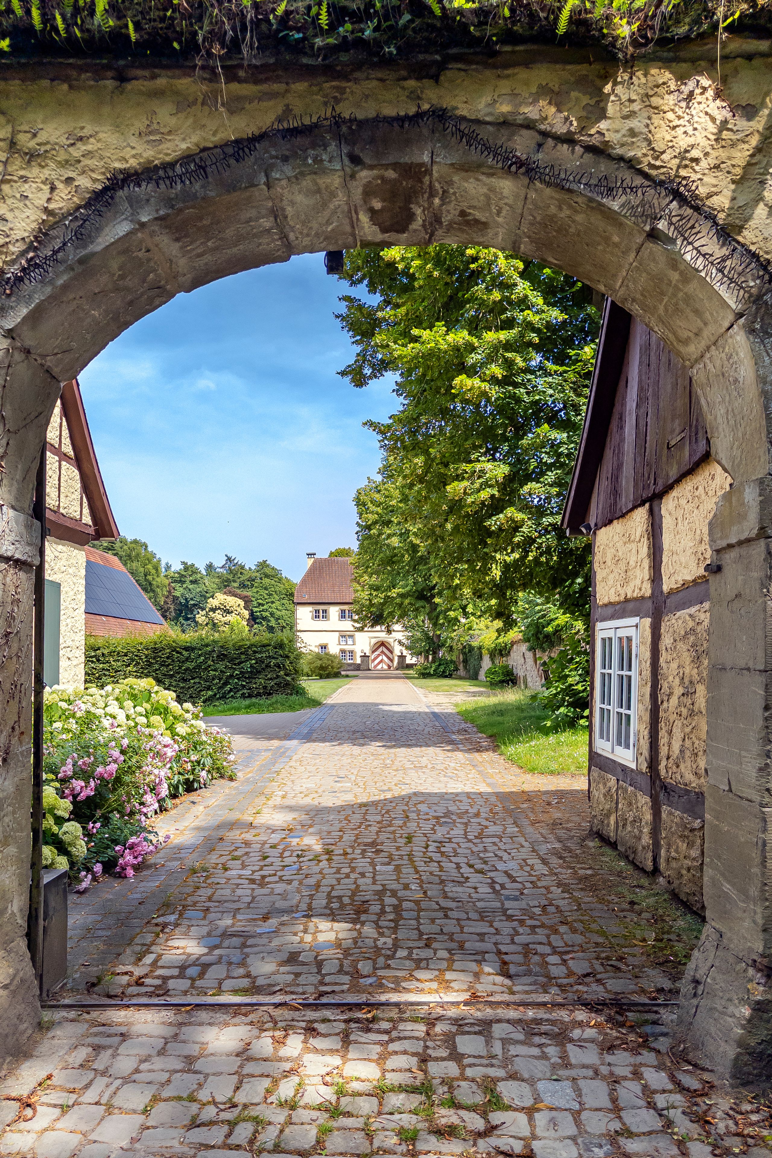 A cobblestone path under an archway leading to a traditional building with a sloped roof.