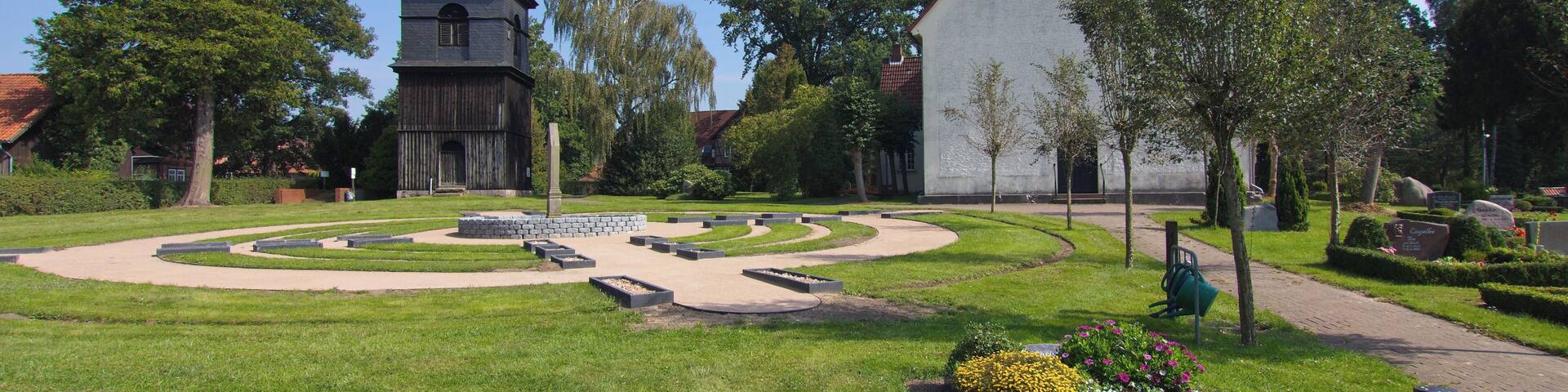 A church with a tower, a white building, and a landscaped garden with flowers and a circular path.