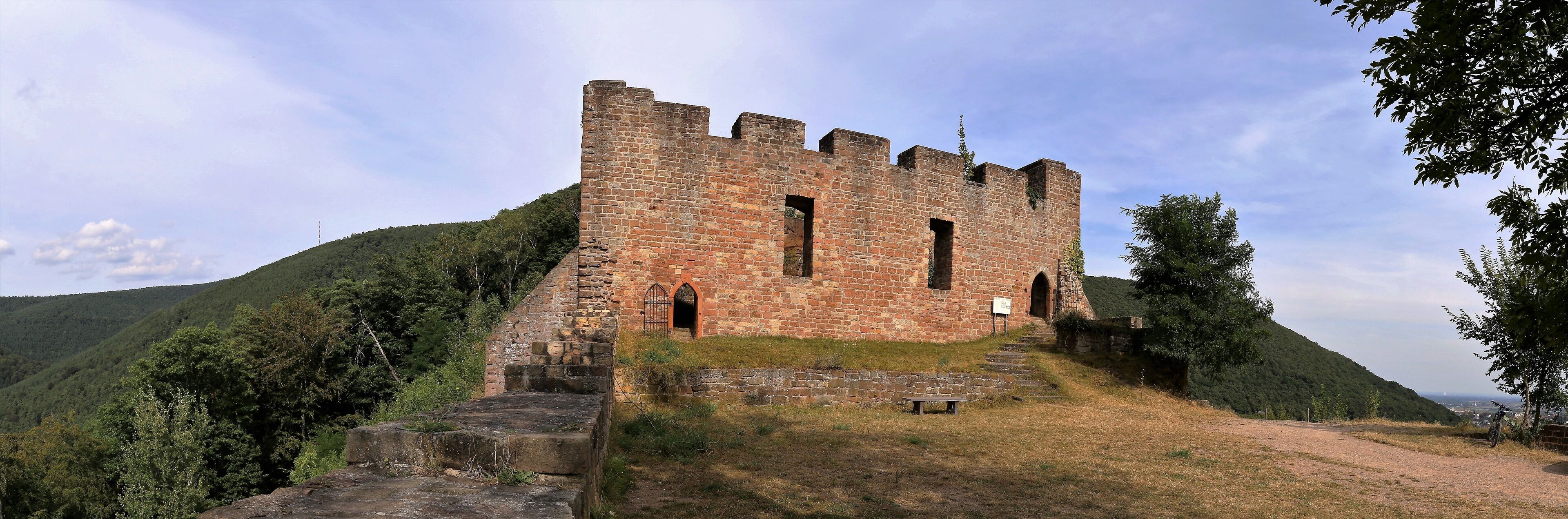 A historic stone castle with multiple towers and arched windows, surrounded by lush green hills and a clear sky.