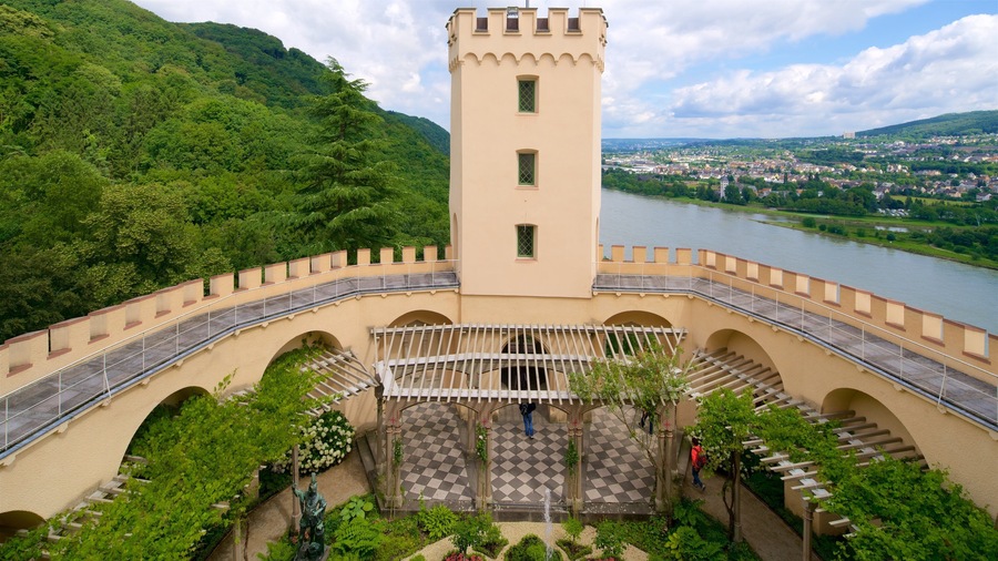 Un château doté d’une tour qui surplombe une rivière, entouré d’une végétation luxuriante et avec une ville visible au loin.