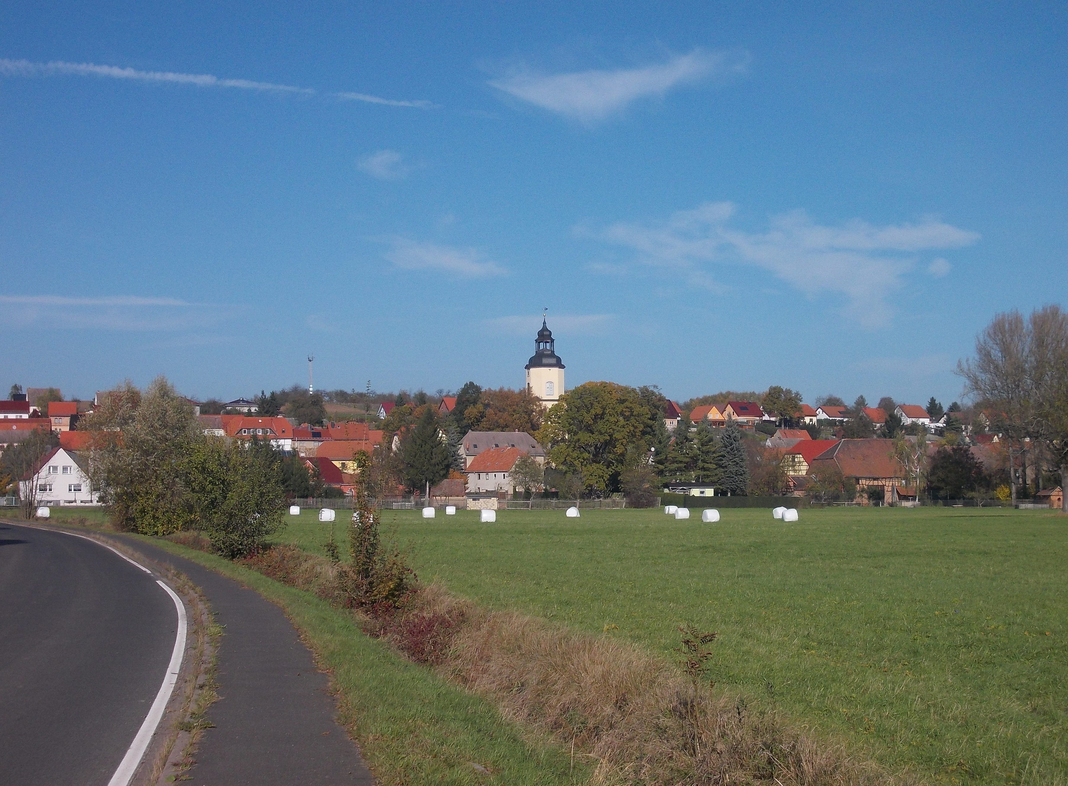 A rural landscape with a church tower, hay bales, and houses.