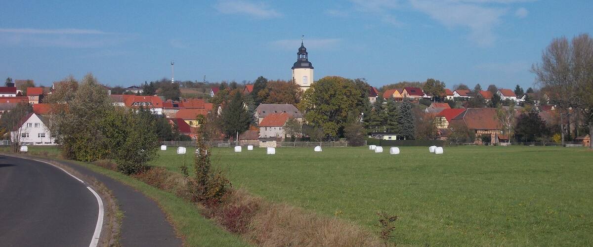 A rural landscape with a church tower, hay bales, and houses.