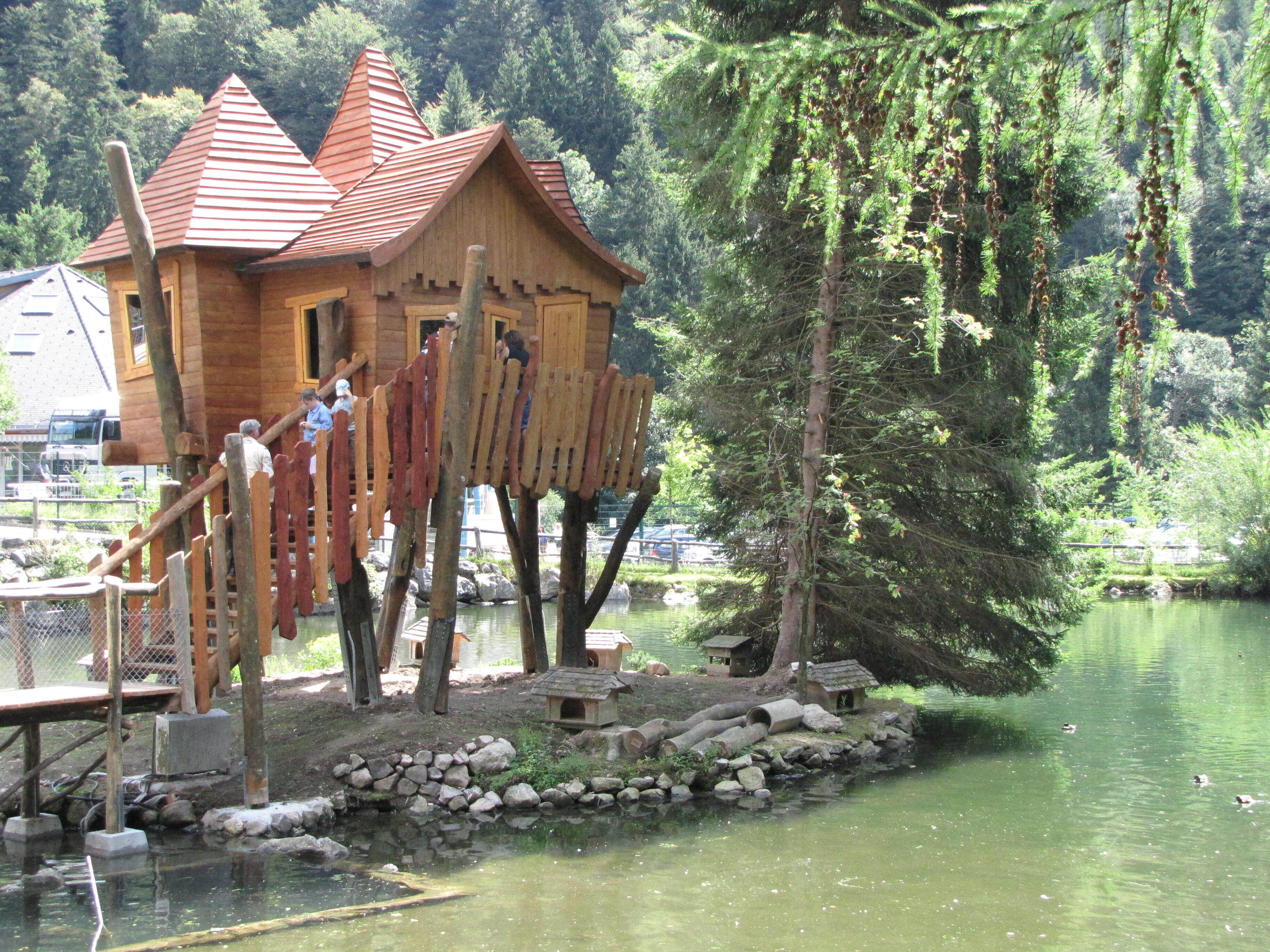 A wooden house with a red roof on stilts by a lake surrounded by trees.